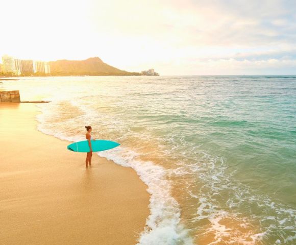 A person stands on a sunlit beach holding a surfboard, gazing at the ocean. The scene conveys tranquility as waves gently lap the shore at sunset.