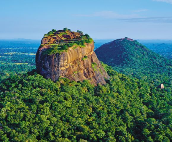 Aerial view of Sigiriya Rock Fortress in Sri Lanka, surrounded by lush green forests under a clear blue sky, evoking a sense of majesty and tranquility.