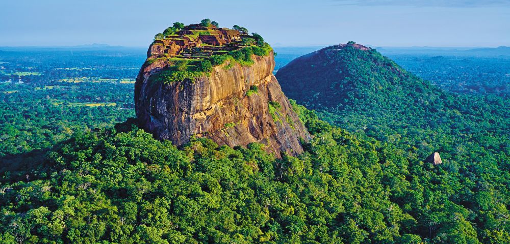 Aerial view of Sigiriya Rock Fortress in Sri Lanka, surrounded by lush green forests under a clear blue sky, evoking a sense of majesty and tranquility.