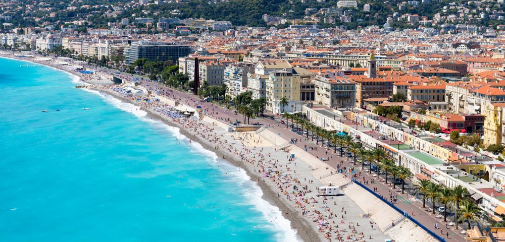 Aerial view of a vibrant beach in Nice, France, with turquoise water, sunbathers on the pebbled shore, a lively promenade, and hills in the background.