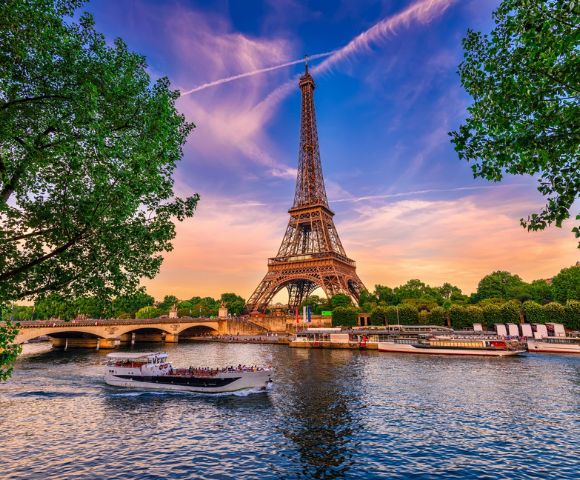 A stunning evening view of the Eiffel Tower from across the Seine River. A tour boat glides by, with vibrant skies and lush trees framing the scene.
