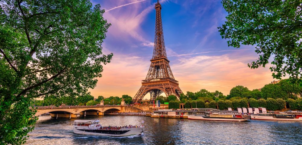 A stunning evening view of the Eiffel Tower from across the Seine River. A tour boat glides by, with vibrant skies and lush trees framing the scene.