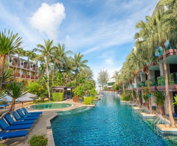 Resort pool scene with blue loungers, flanked by lush palm trees and modern buildings. Bright, sunny day with a serene and inviting atmosphere.
