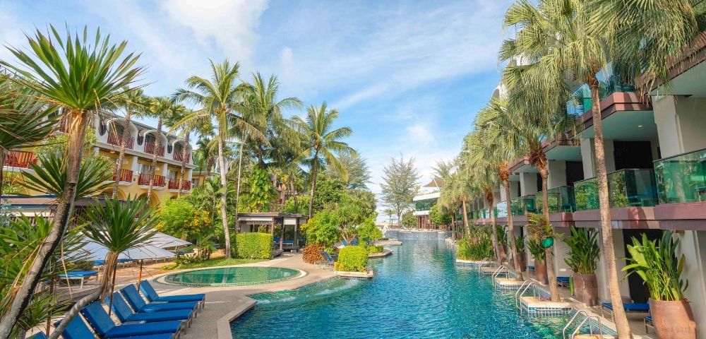 Resort pool scene with blue loungers, flanked by lush palm trees and modern buildings. Bright, sunny day with a serene and inviting atmosphere.