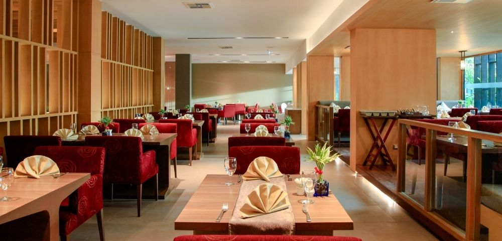 Cozy restaurant interior featuring red upholstered chairs, neatly set tables with folded napkins, wooden accents, dim lighting, and a welcoming ambiance.