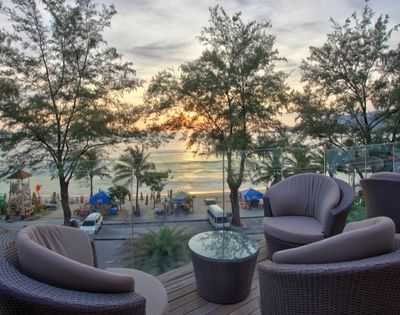 Cozy patio with wicker chairs and a glass table overlooks a beach at sunset. Palm trees and gentle waves create a serene, relaxing atmosphere.