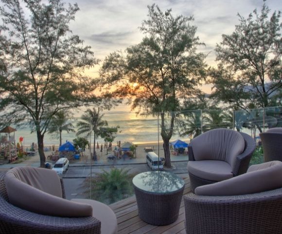 Cozy patio with wicker chairs and a glass table overlooks a beach at sunset. Palm trees and gentle waves create a serene, relaxing atmosphere.