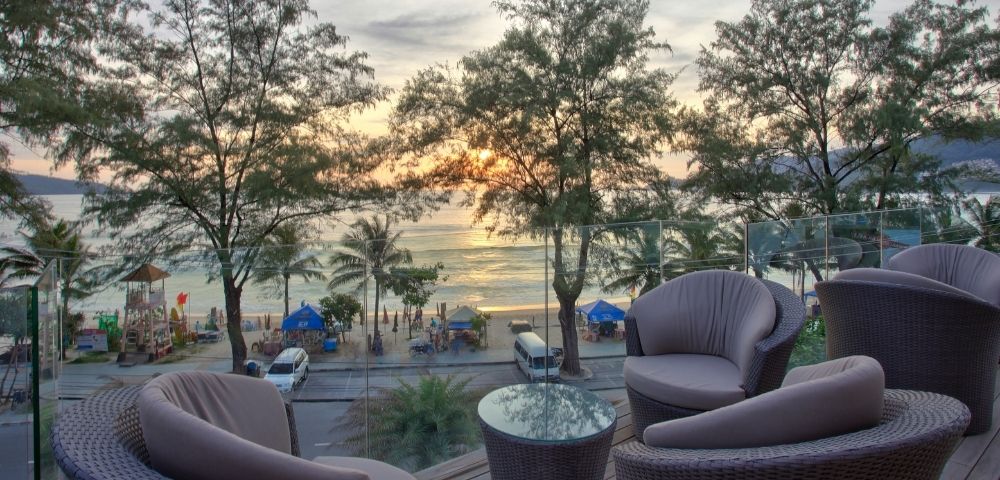 Cozy patio with wicker chairs and a glass table overlooks a beach at sunset. Palm trees and gentle waves create a serene, relaxing atmosphere.