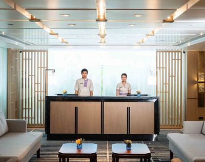 Two hotel receptionists stand behind a modern front desk in a bright, elegant lobby. The space features wooden accents, sofas, and warm lighting.