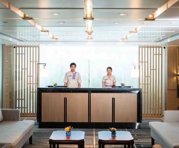 Two hotel receptionists stand behind a modern front desk in a bright, elegant lobby. The space features wooden accents, sofas, and warm lighting.