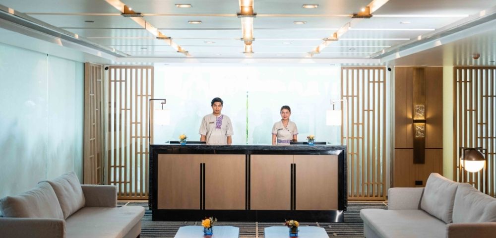 Two hotel receptionists stand behind a modern front desk in a bright, elegant lobby. The space features wooden accents, sofas, and warm lighting.