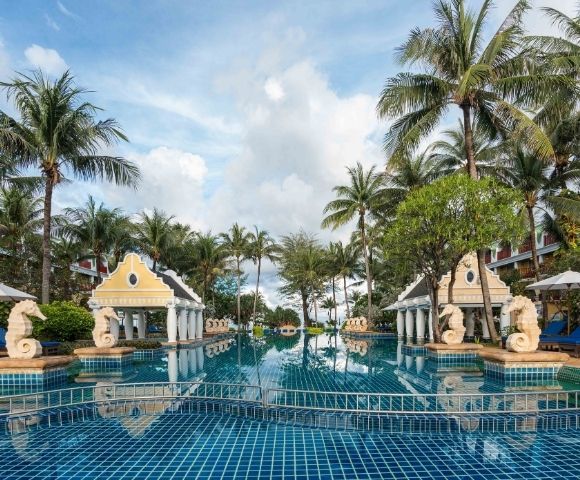 A luxurious resort pool framed by palm trees, ornate columns, and seahorse statues. The serene water reflects the partly cloudy sky above.