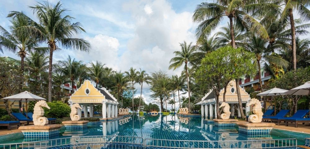 A luxurious resort pool framed by palm trees, ornate columns, and seahorse statues. The serene water reflects the partly cloudy sky above.