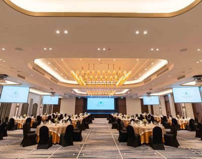 Elegant banquet hall with a large chandelier, round tables covered in gold and black, and projection screens. The atmosphere is sophisticated and formal.