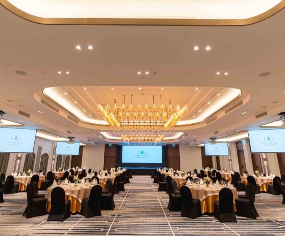 Elegant banquet hall with a large chandelier, round tables covered in gold and black, and projection screens. The atmosphere is sophisticated and formal.