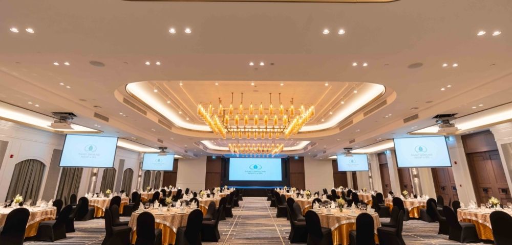 Elegant banquet hall with a large chandelier, round tables covered in gold and black, and projection screens. The atmosphere is sophisticated and formal.