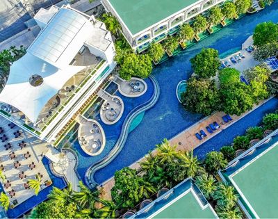 Aerial view of a luxurious hotel with a curving swimming pool surrounded by lush greenery, lounge chairs, and a modern building with a unique roof design.