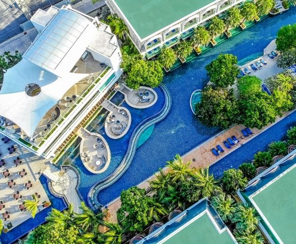 Aerial view of a luxurious hotel with a curving swimming pool surrounded by lush greenery, lounge chairs, and a modern building with a unique roof design.