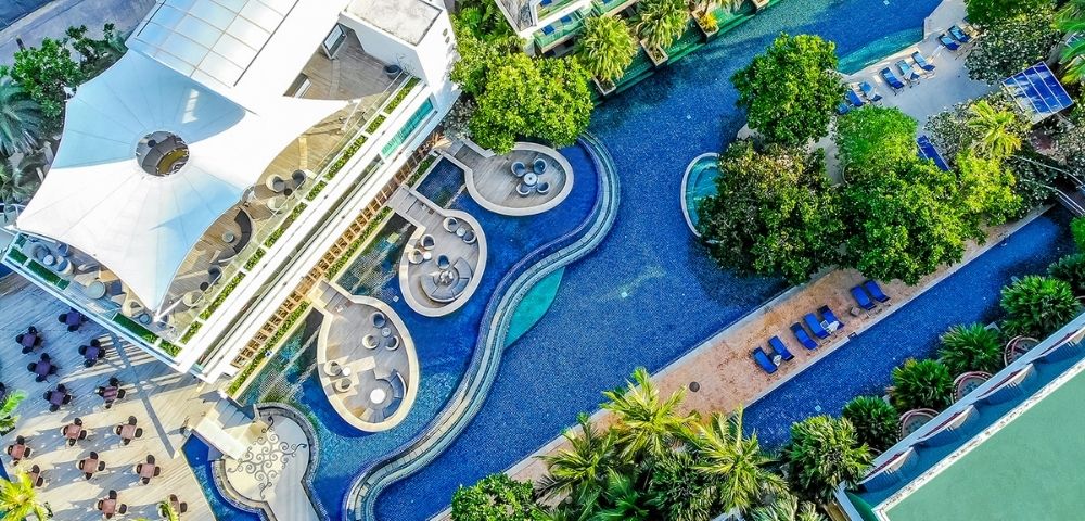 Aerial view of a luxurious hotel with a curving swimming pool surrounded by lush greenery, lounge chairs, and a modern building with a unique roof design.