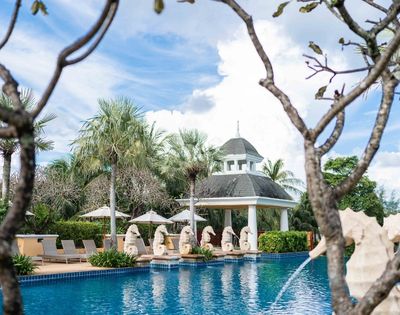 Serene pool area with clear blue water, surrounded by palm trees and chaise lounges. Statues line the poolside, with a gazebo in the background under a bright sky.