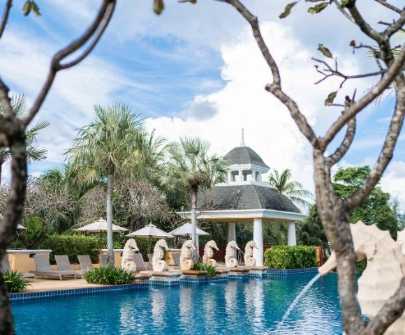 Serene pool area with clear blue water, surrounded by palm trees and chaise lounges. Statues line the poolside, with a gazebo in the background under a bright sky.