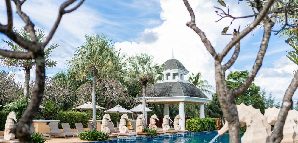 Serene pool area with clear blue water, surrounded by palm trees and chaise lounges. Statues line the poolside, with a gazebo in the background under a bright sky.