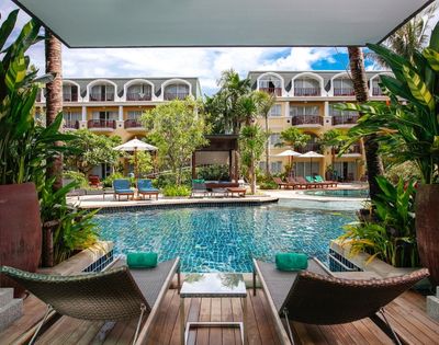 Two lounge chairs with teal towels face a serene pool surrounded by lush greenery and a hotel building. The scene conveys relaxation and luxury.