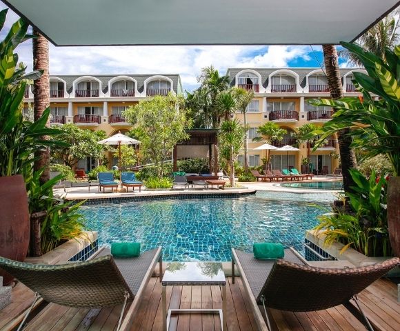 Two lounge chairs with teal towels face a serene pool surrounded by lush greenery and a hotel building. The scene conveys relaxation and luxury.