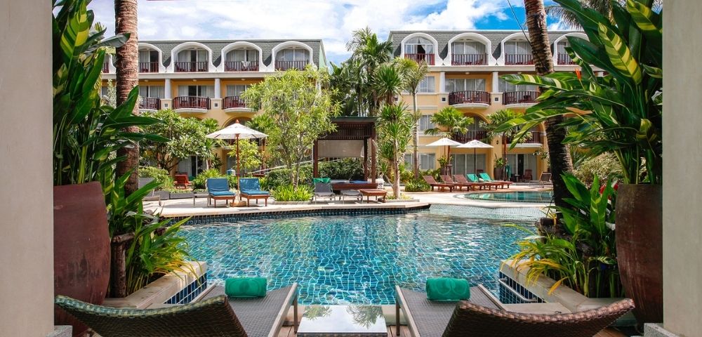 Two lounge chairs with teal towels face a serene pool surrounded by lush greenery and a hotel building. The scene conveys relaxation and luxury.