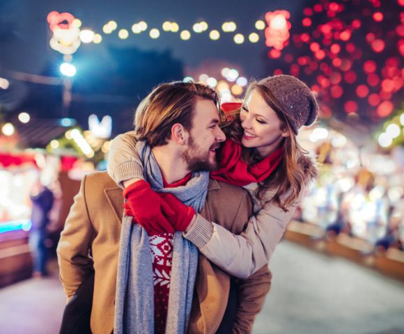 A couple enjoys a winter market, with the woman playfully hugging the man from behind. They smile amid colorful lights, conveying warmth and joy.