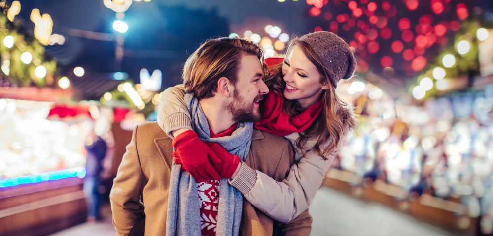 A couple enjoys a winter market, with the woman playfully hugging the man from behind. They smile amid colorful lights, conveying warmth and joy.