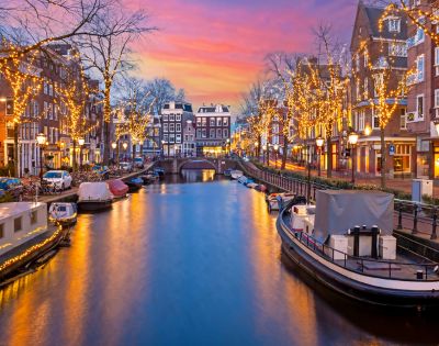 A serene canal in Amsterdam at sunset, adorned with twinkling fairy lights on trees. Boats line the water, reflecting the vibrant pink and orange sky.