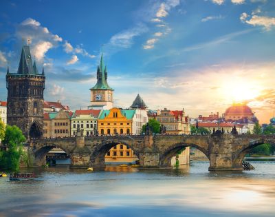 Sunlit view of Charles Bridge and historic towers in Prague. The scene features vibrant architecture, reflected in the Vltava River, under a bright, clear sky.
