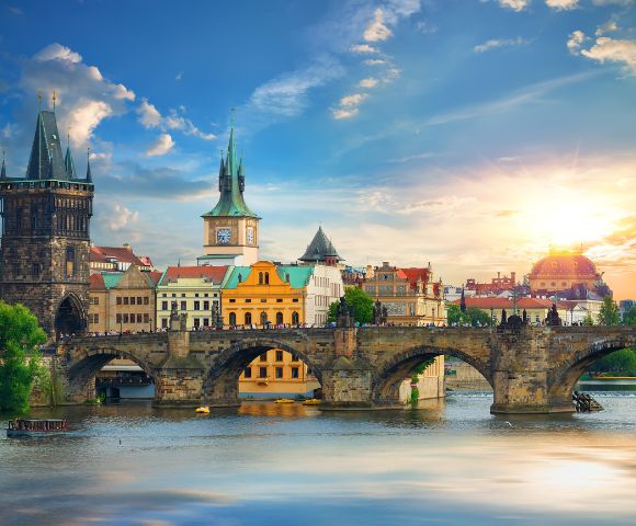Sunlit view of Charles Bridge and historic towers in Prague. The scene features vibrant architecture, reflected in the Vltava River, under a bright, clear sky.