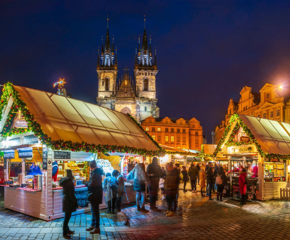 Festive Christmas market at night with illuminated booths decorated with garlands in a historic square. People shop under a deep blue sky, creating a warm, lively atmosphere.