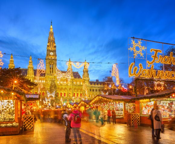 Festive Christmas market scene with glowing lights and stalls in front of a lit-up building under a twilight sky. People walk, creating a warm, lively atmosphere.
