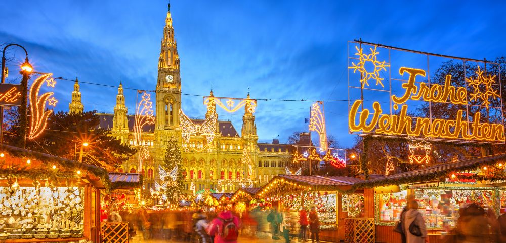 Festive Christmas market scene with glowing lights and stalls in front of a lit-up building under a twilight sky. People walk, creating a warm, lively atmosphere.