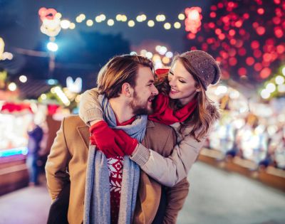 A couple enjoys a winter market, with the woman playfully hugging the man from behind. They smile amid colorful lights, conveying warmth and joy.