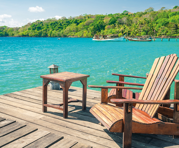 A wooden deck overlooks clear blue water with lush green trees in the background. Two wooden chairs and a small table create a tranquil, relaxing scene.