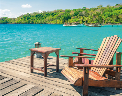 A wooden deck overlooks clear blue water with lush green trees in the background. Two wooden chairs and a small table create a tranquil, relaxing scene.