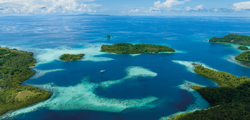 Aerial view of a tropical seascape with lush green islands surrounded by turquoise water and coral reefs. A small boat is visible, evoking serenity.