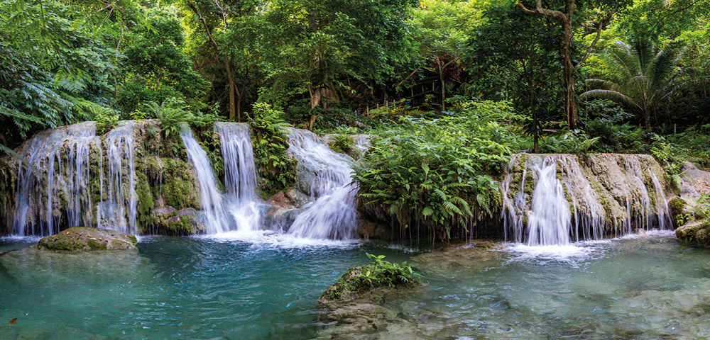 A serene waterfall cascades into turquoise pools surrounded by lush greenery. Sunlight filters through trees, creating a tranquil and refreshing atmosphere.