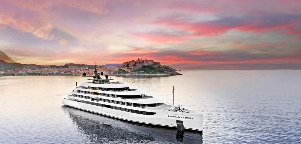 The luxury yacht Emerald Azzurra cruising on tranquil seas at sunset, with a Mediterranean coastal town and pastel sky in the background.