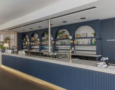 Sleek modern bar with navy blue accents, shelves stocked with bottles, potted plants, and framed art. Bright, inviting atmosphere.