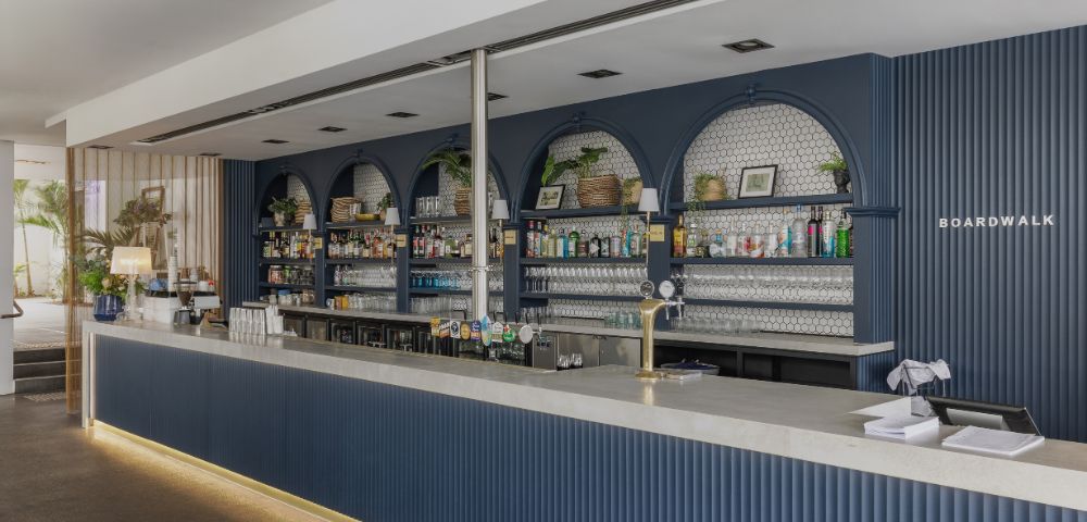 Sleek modern bar with navy blue accents, shelves stocked with bottles, potted plants, and framed art. Bright, inviting atmosphere.