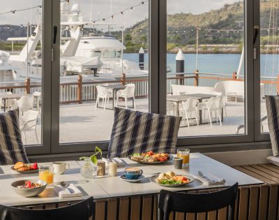 Coastal dining scene with a table set for breakfast, featuring fruit, pastries, and juice. Large windows offer a view of yachts and a serene marina.