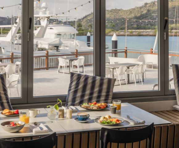 Coastal dining scene with a table set for breakfast, featuring fruit, pastries, and juice. Large windows offer a view of yachts and a serene marina.