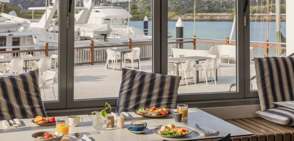 Coastal dining scene with a table set for breakfast, featuring fruit, pastries, and juice. Large windows offer a view of yachts and a serene marina.