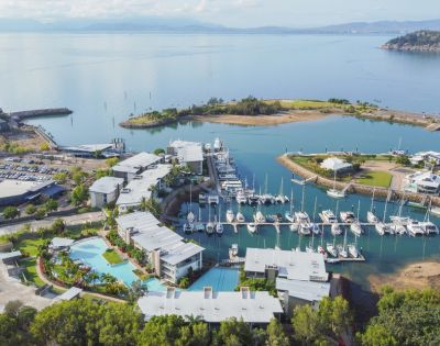 Aerial view of a marina with several boats docked. Modern buildings and pools in the foreground, surrounded by greenery. Calm sea and distant hills.