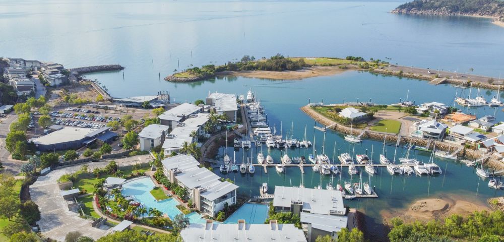 Aerial view of a marina with several boats docked. Modern buildings and pools in the foreground, surrounded by greenery. Calm sea and distant hills.